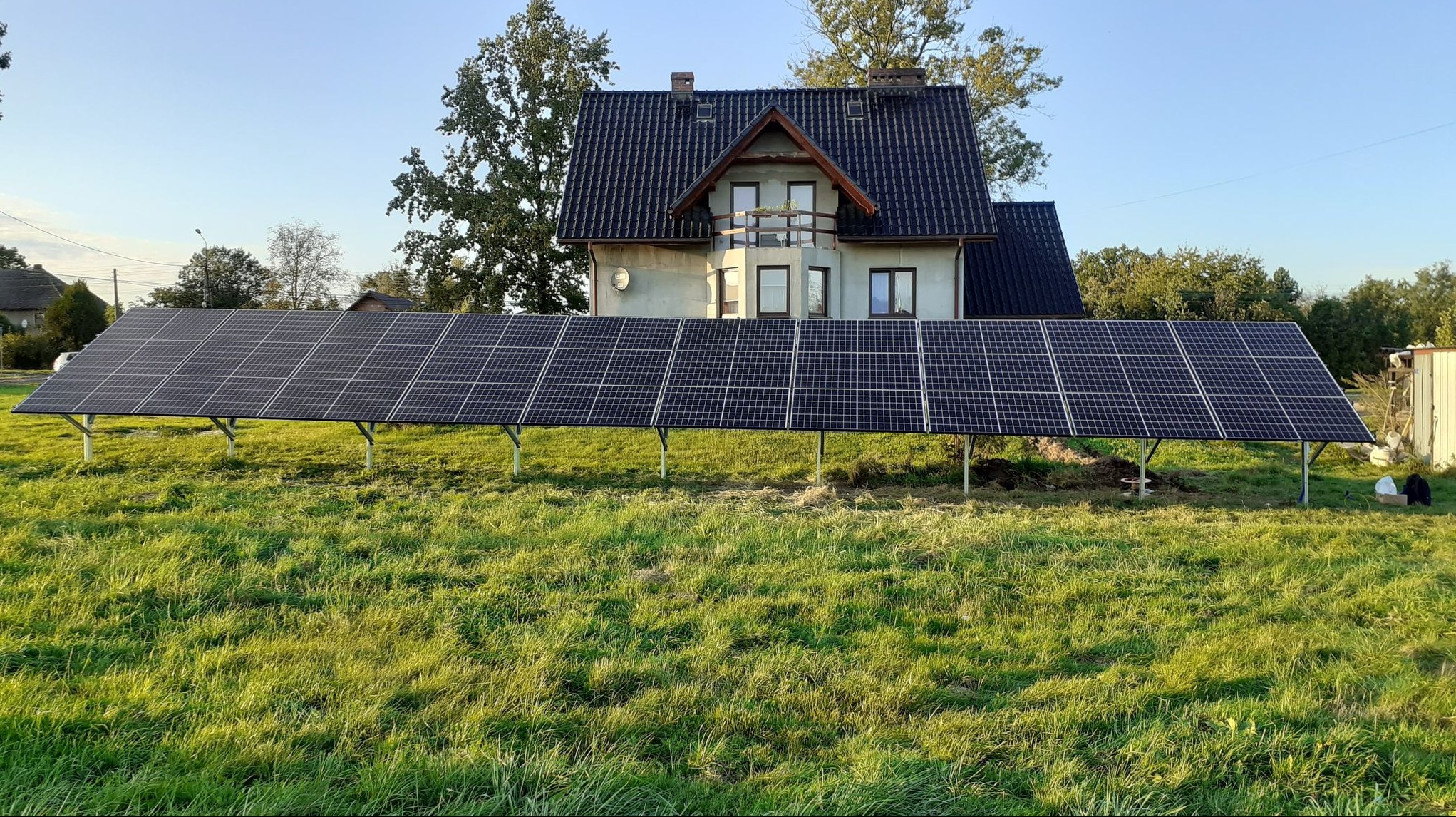 Rząd paneli słonecznych na metalowych wspornikach, ustawiony na trawiastym terenie przed domem jednorodzinnym z ciemnym dachem, w słoneczny dzień.