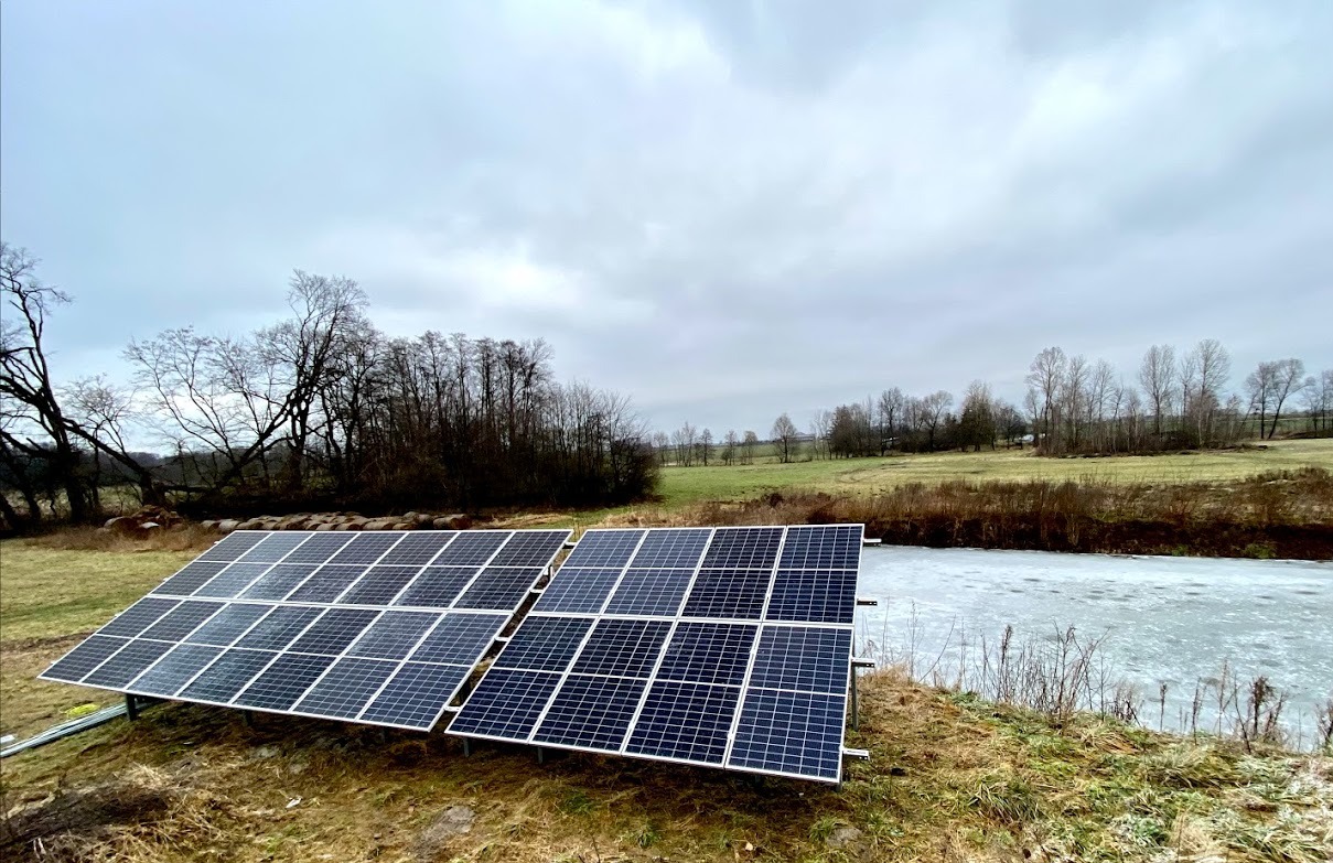 Dwa rzędy paneli słonecznych zainstalowane na łące w pobliżu częściowo zamarzniętego zbiornika wodnego, w tle drzewa bez liści i pochmurne niebo.