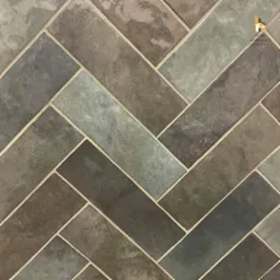 Ułożona w jodełkę mozaika z płytek w odcieniach szarości i brązu z jasną fugą, widoczne logo firmy w prawym górnym rogu.