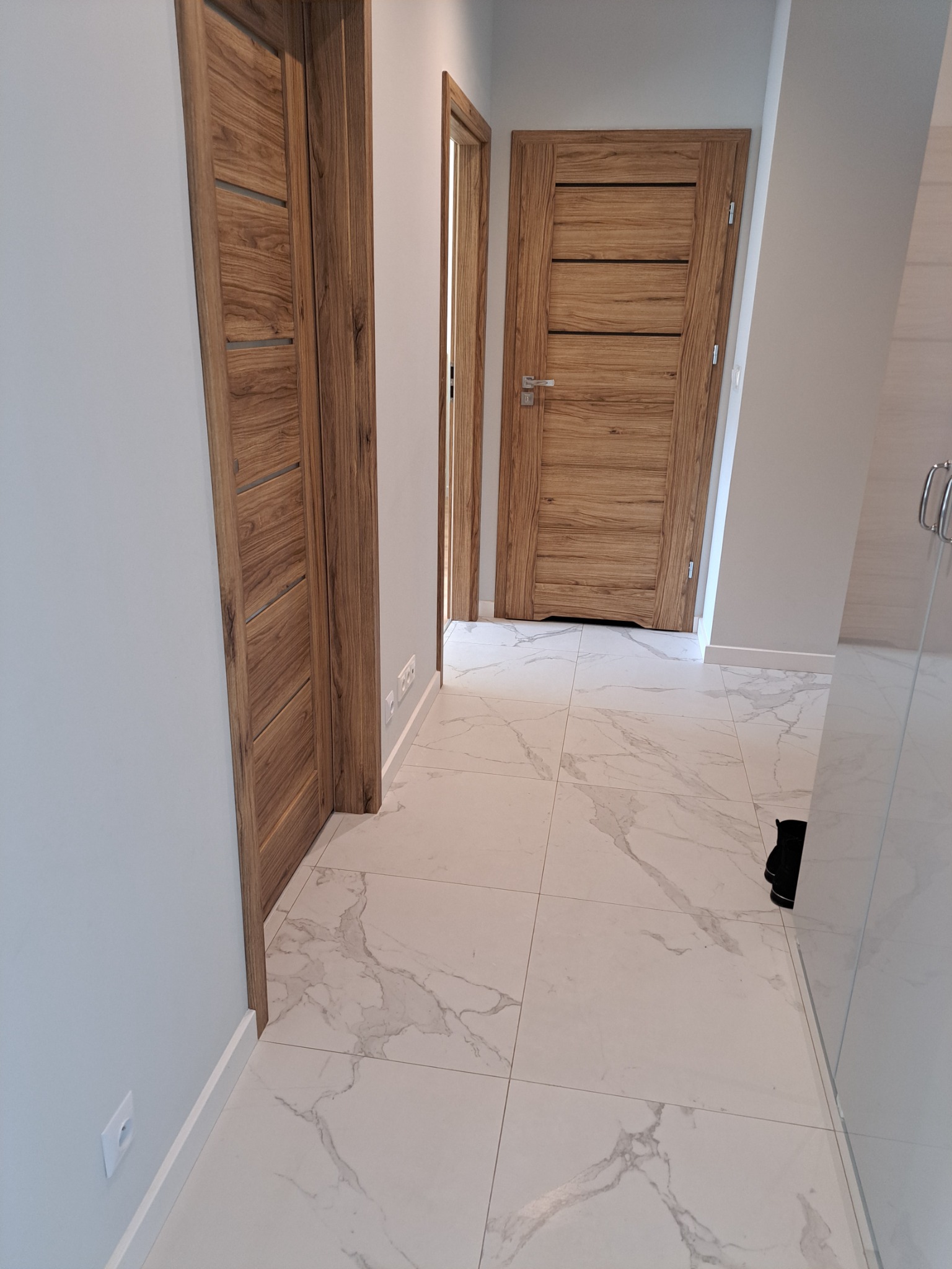 Wnętrze korytarza z trzema drewnianymi drzwiami o nowoczesnym designie, białe ściany i marmurowa podłoga z szarymi żyłami, widoczne gniazdka elektryczne przy listwach przypodłogowych.