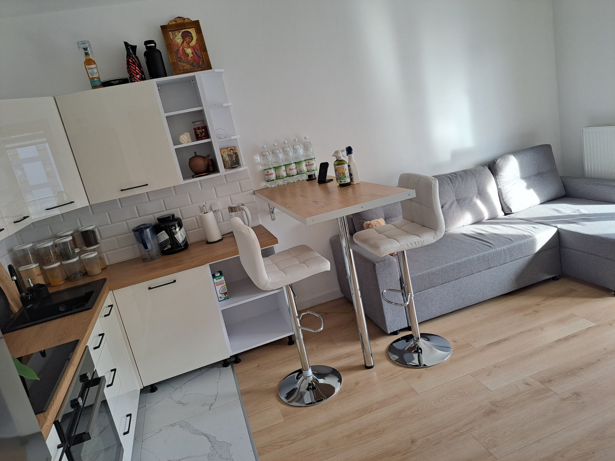 Jasne wnętrze mieszkania z aneksem kuchennym, wysokimi krzesłami barowymi i szarą narożną sofą, widok na podłogę z paneli.