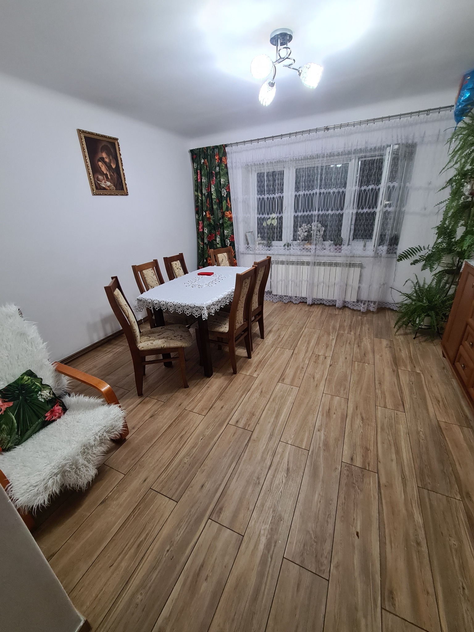 Jasne wnętrze pokoju z drewnianą podłogą, stołem nakrytym białym obrusem z koronką, krzesłami z tapicerką w kwiaty, białym fotelem z poduszką w kwiaty i zielonymi roślinami w doniczkach przy oknie.