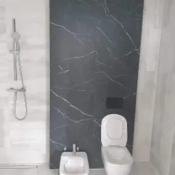 Wykończona łazienka z czarną ścianą z efektem marmuru, biała armatura: toaleta i bidet na kartonie ochronnym, srebrna bateria prysznicowa, jasne płytki podłogowe i ścienne.