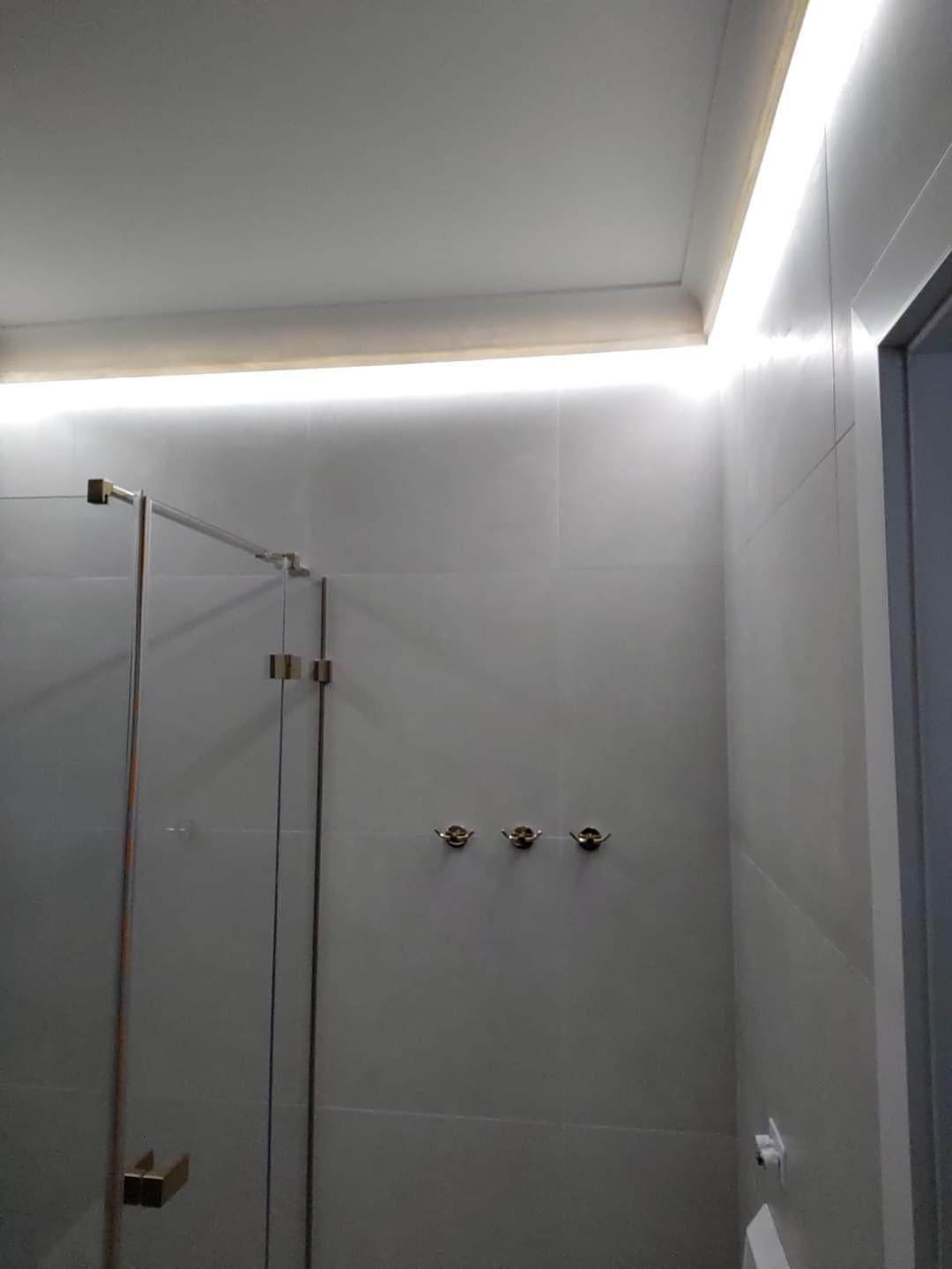 Wykończona łazienka z kabiną prysznicową ze szklanymi drzwiami, oświetleniem LED podsufitowym i trzema ozdobnymi wieszakami na ścianie wyłożonej jasnymi płytkami.