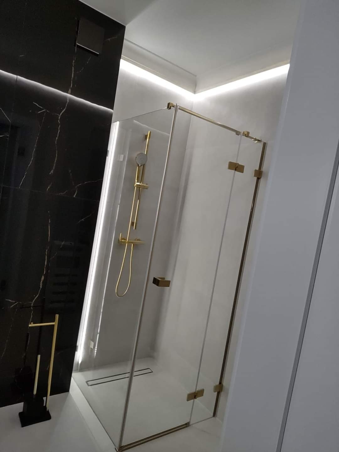 Nowoczesna kabina prysznicowa z złotymi okuciami, czarną armaturą i czarnym marmurem w tle, oświetlenie LED w suficie.