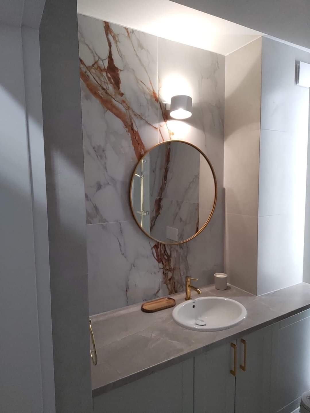 Nowoczesna łazienka z marmurowymi płytkami, okrągłym lustrem w złotej ramie, białą umywalką i złotą baterią, oświetlona kinkietem.