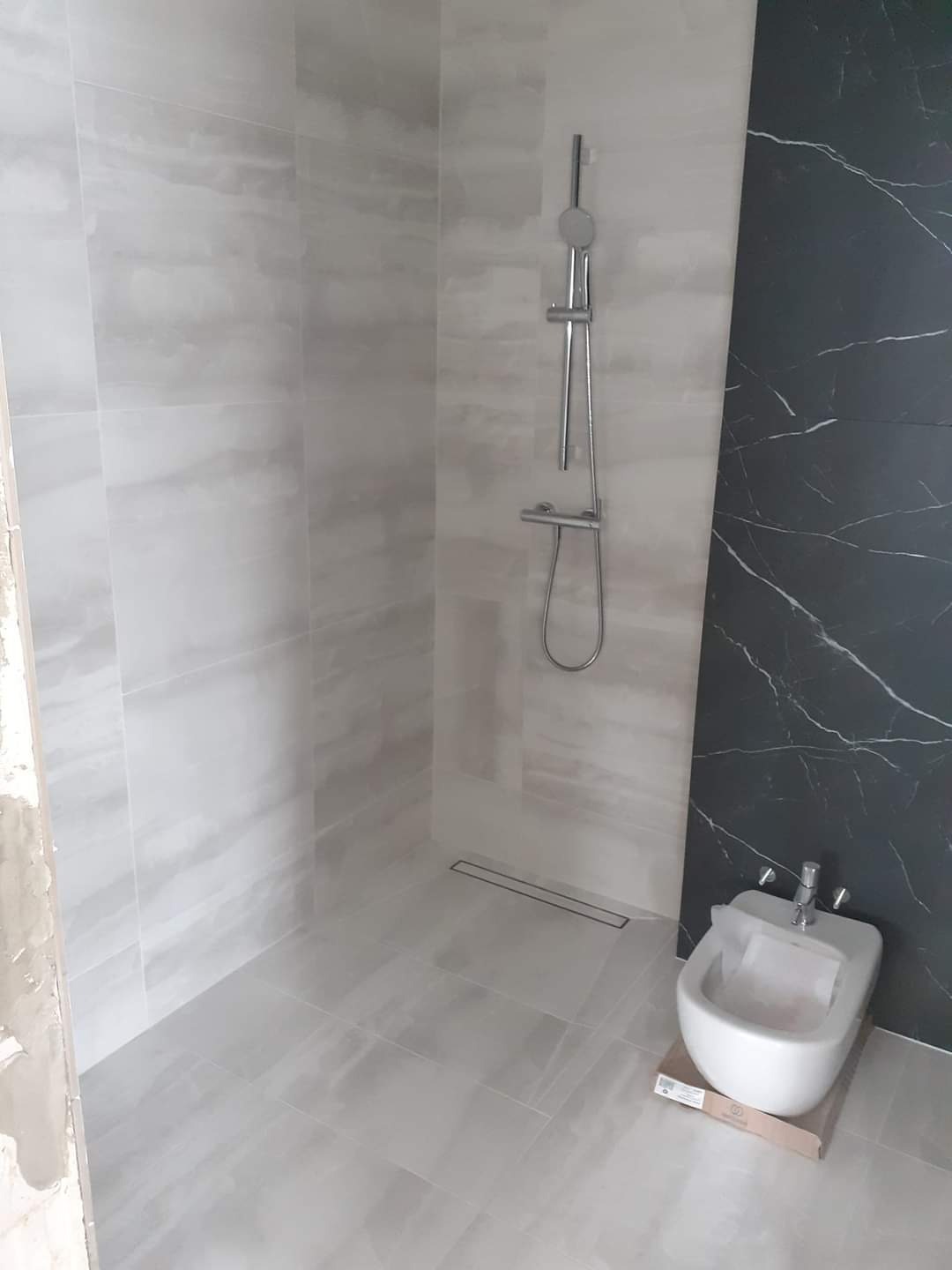 Wnętrze łazienki w trakcie remontu, widoczne jasnoszare płytki imitujące marmur na ścianach i podłodze, chromowana armatura prysznicowa oraz biały bidet stojący na kartonie, fragment surowej ściany...