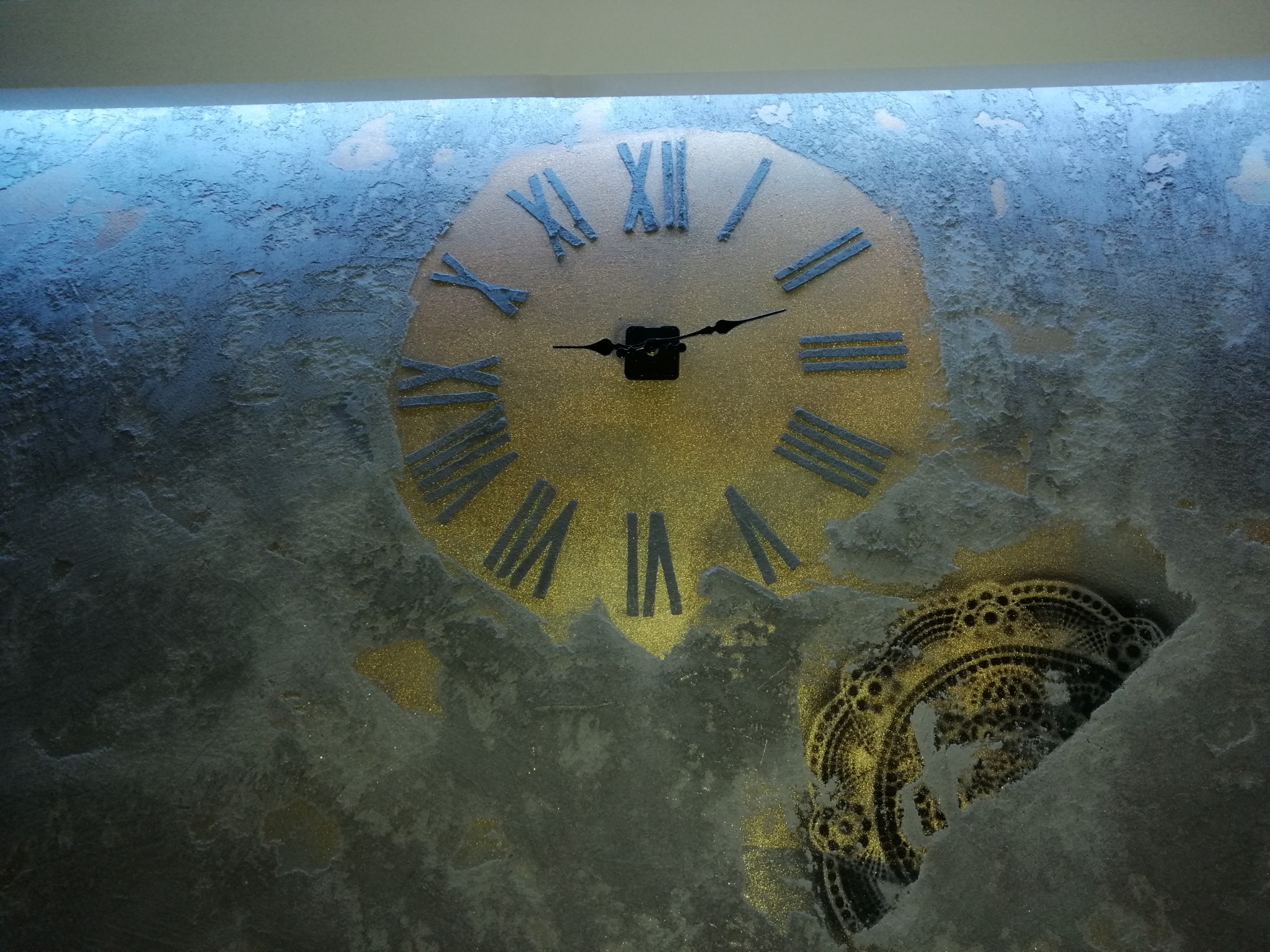 Dekoracyjna ściana z efektem gładzi, zdobiona dużym zegarem w stylu vintage i ornamentem w kolorze złota.