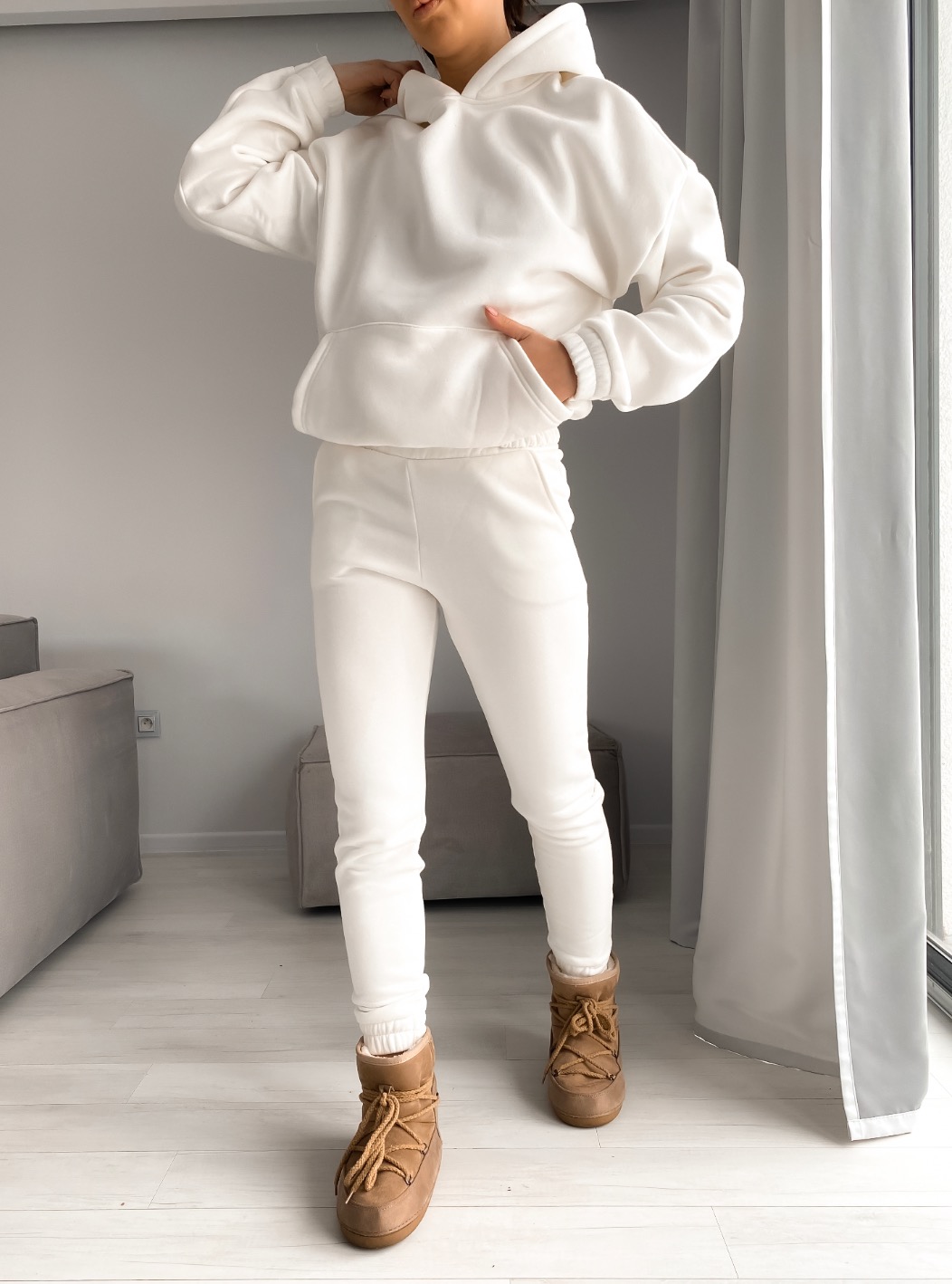 Kobieta prezentuje biały, dresowy komplet składający się z bluzy z kapturem i spodni dresowych, uzupełniony brązowymi butami typu moon boot przy oknie.