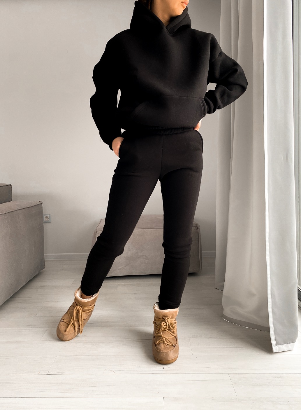 Kobieta pozuje w czarnym, dresowym komplecie z kapturem i brązowych śniegowcach w minimalistycznym wnętrzu z szarymi meblami i białymi zasłonami.