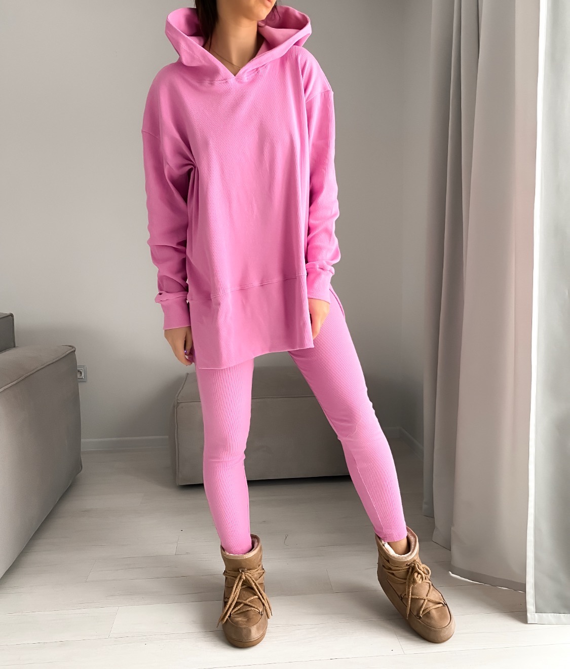 Kobieta prezentuje różowy, prążkowany komplet dresowy składający się z bluzy z kapturem i legginsów, uzupełniony brązowymi butami typu śniegowce.