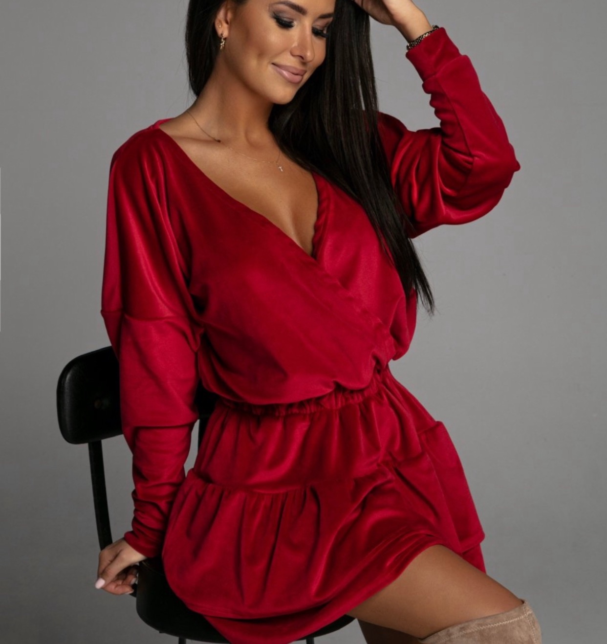 Uśmiechnięta brunetka w czerwonej, welurowej sukience z dekoltem w kształcie litery V, siedząca na czarnym krześle, z ręką uniesioną do włosów, na szarym tle.