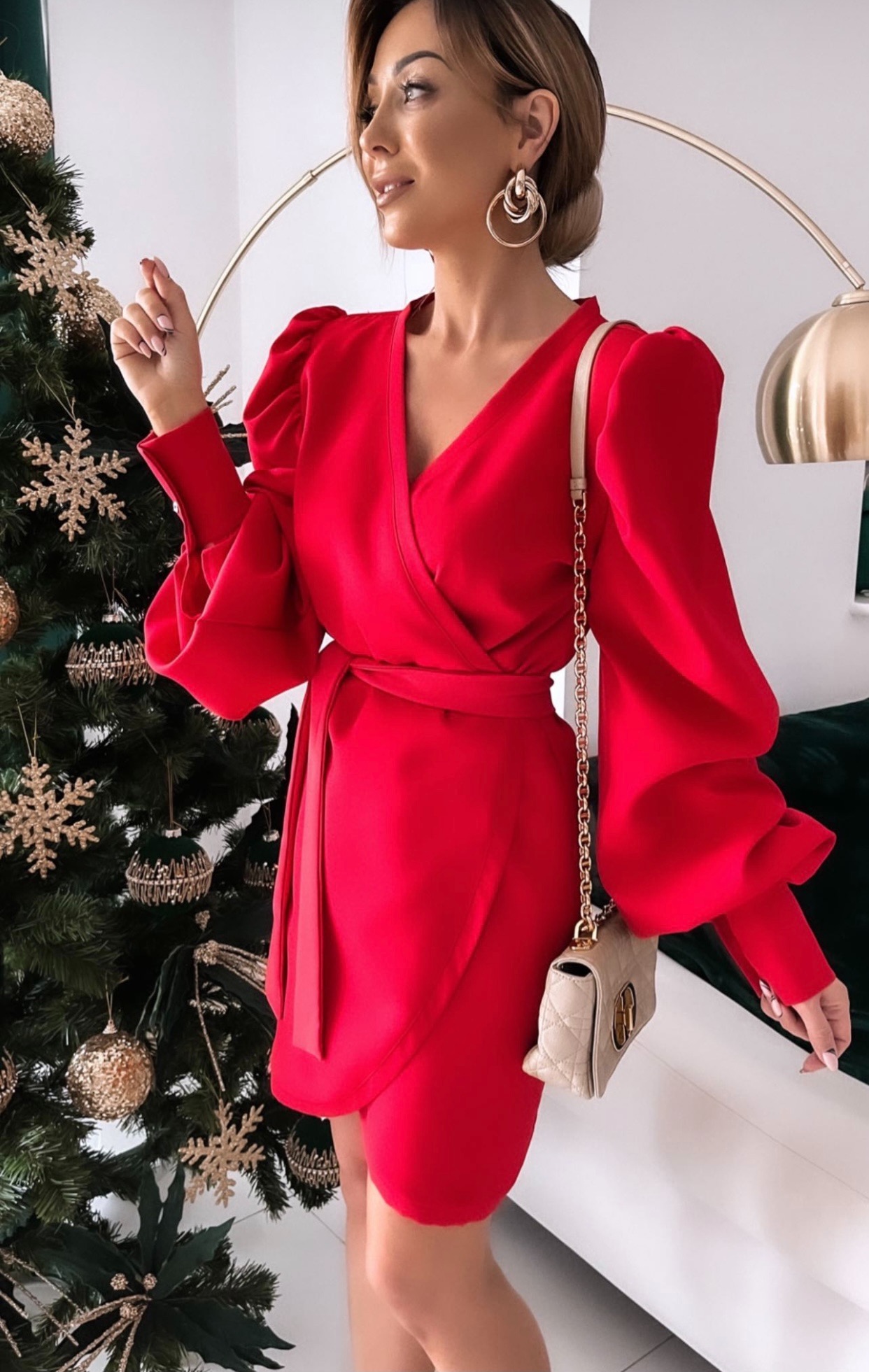 Kobieta w czerwonej, kopertowej sukience z bufiastymi rękawami, pozująca obok choinki z ozdobami w odcieniach złota, z beżową torebką na łańcuszku.