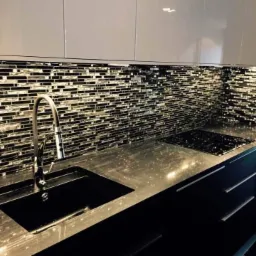 Nowoczesna kuchnia w Gnieźnie z błyszczącą, czarno-srebrną mozaiką na ścianie nad blatem, czarnym zlewem i płytą grzewczą oraz białymi szafkami wiszącymi.