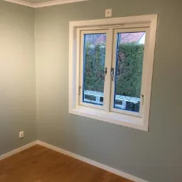 Pomalowane na jasny odcień zieleni ściany pokoju z drewnianą podłogą i białymi listwami przypodłogowymi, widoczne okno z białymi ramami, za oknem zieleń i drabina oparta o ścianę.