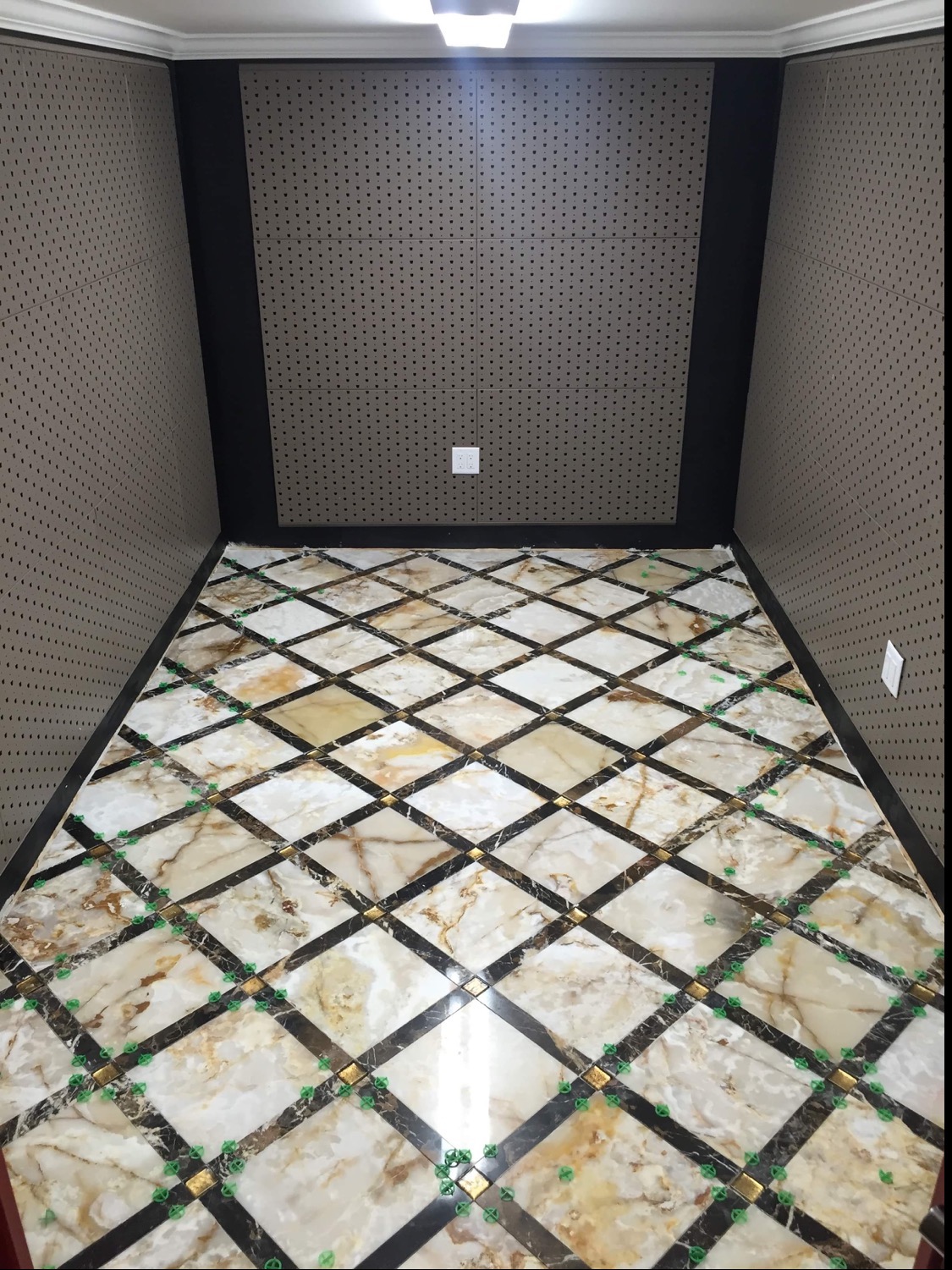 Podłoga wyłożona marmurowymi płytkami w romby z czarnymi wstawkami, widoczne plastikowe krzyżyki dystansowe, ściany wykończone panelami z perforacją, oświetlenie sufitowe.
