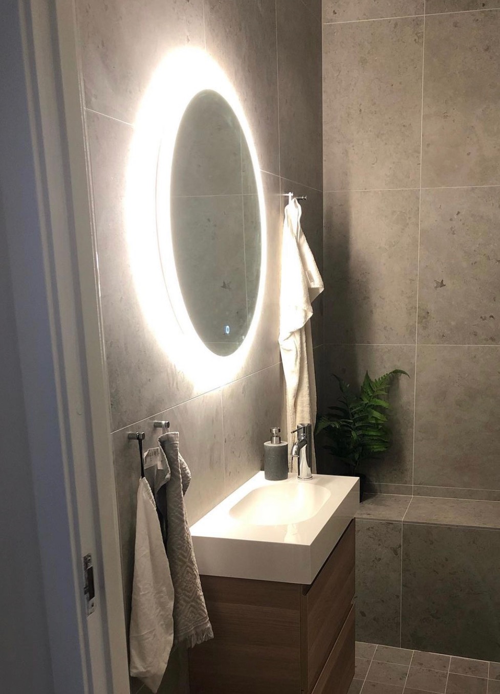 Nowoczesna łazienka z szarymi płytkami, owalnym lustrem podświetlanym LED, białą umywalką nablatową na drewnianej szafce oraz rośliną paproci w rogu.