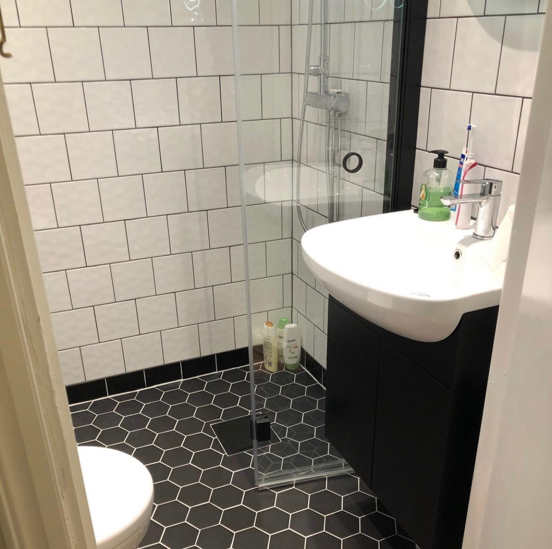 Wykończona łazienka z heksagonalną czarno-białą podłogą, białe kafelki na ścianach, kabina prysznicowa ze szklaną ścianką, biała umywalka z czarną szafką, widoczne kosmetyki i akcesoria łazienkowe.