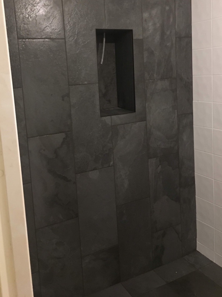 Ściana kabiny prysznicowej wyłożona ciemnoszarymi płytkami imitującymi kamień, z wnęką na kosmetyki, sąsiadująca z fragmentem ściany wyłożonej białymi, kwadratowymi płytkami.