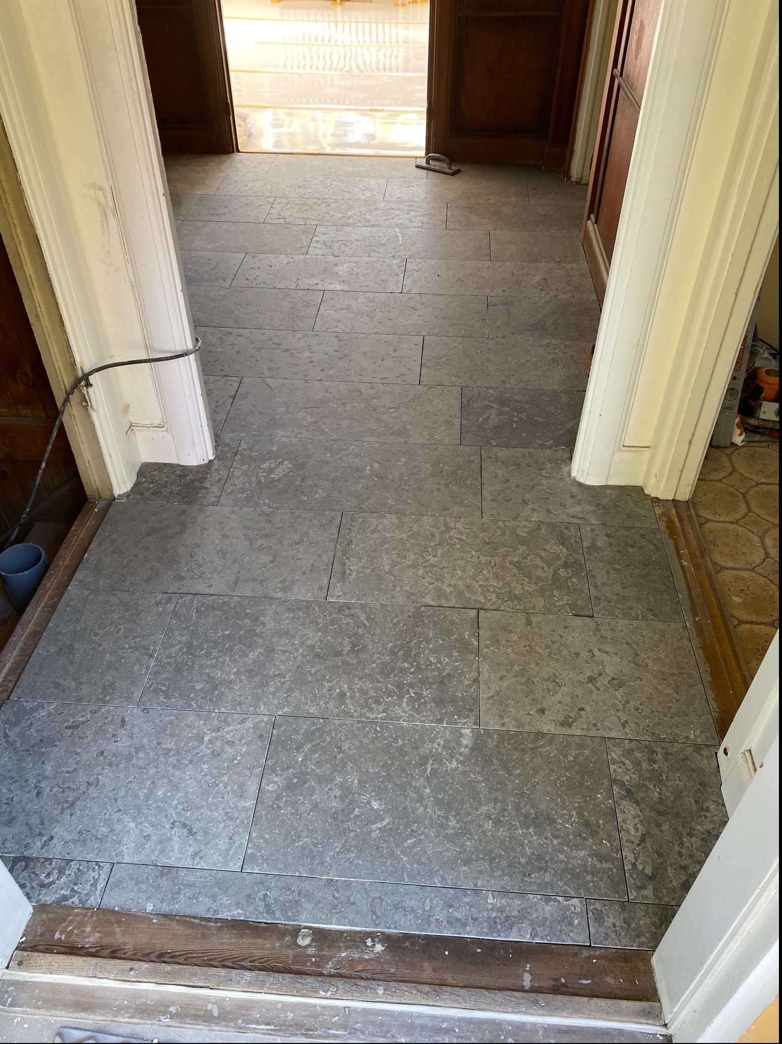Ułożona szara kamienna posadzka w korytarzu, widok przez futrynę z drewnianym progiem, z widocznymi narzędziami w tle i niezakrytą podłogą w sąsiednim pomieszczeniu.
