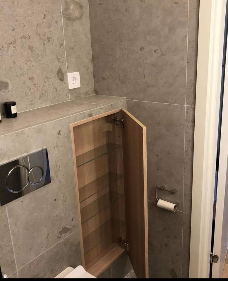 Szara łazienka z otwartą szafką wnękową z drewnianymi półkami, widoczne płytki imitujące kamień, przycisk spłuczki, gniazdko elektryczne i uchwyt na papier toaletowy.