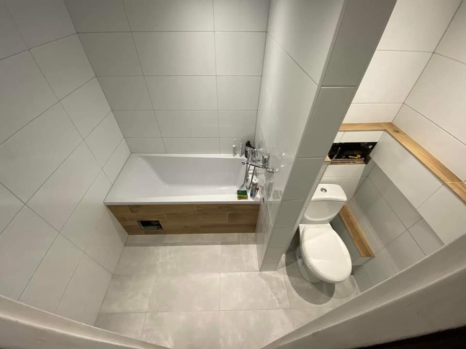 Wnętrze łazienki po remoncie: białe płytki na ścianach i podłodze, zabudowana wanna z drewnopodobnym frontem, toaleta w rogu, drewniane wykończenie wnęki.