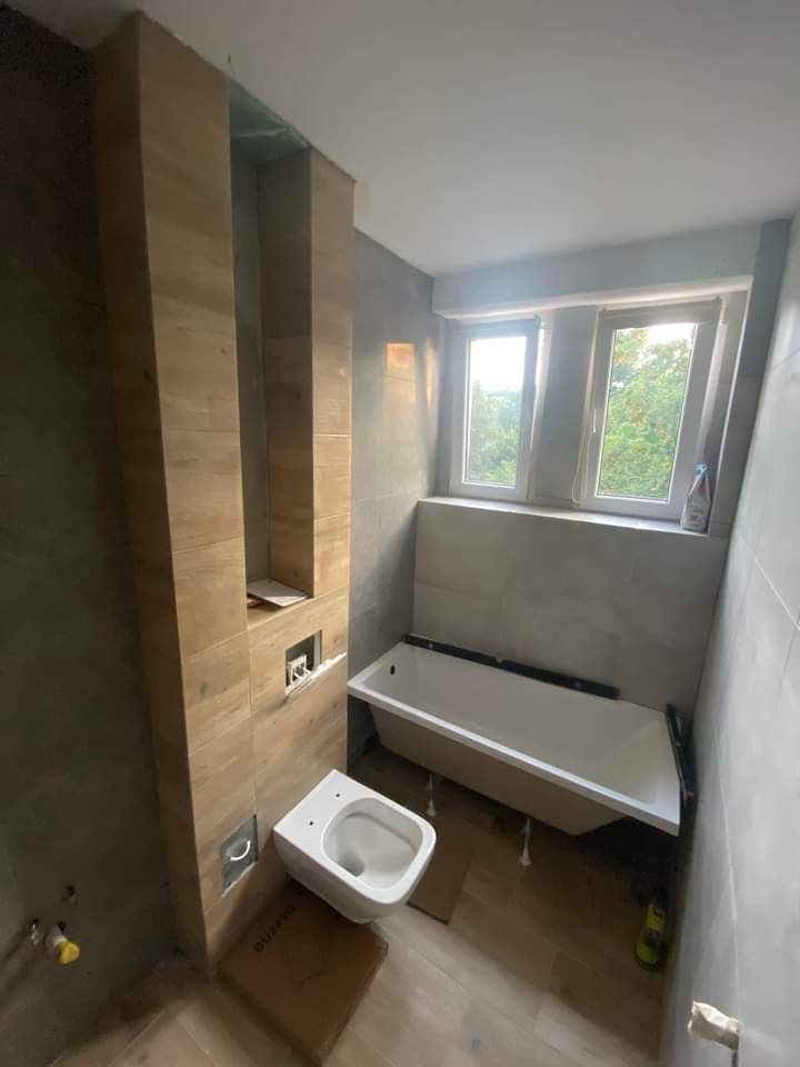 Wnętrze łazienki w trakcie wykończenia: biała wanna, podwieszana toaleta na kartonie, pionowy fragment ściany wyłożony płytkami imitującymi drewno, szare płytki na pozostałych ścianach, okno...