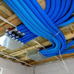 Niebieskie rury wentylacyjne przymocowane do drewnianego stropu z metalowymi skrzynkami rozdzielczymi i białymi kablami, widoczne bloczki z białej cegły i okno.