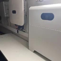 Biała bateria magazynująca energię Huawei obok białej skrzynki elektrycznej, zamontowane na ścianie w pomieszczeniu gospodarczym z widocznymi kablami.