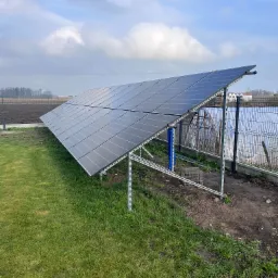 Panel słoneczny ustawiony na metalowej konstrukcji na trawiastym terenie, widoczny fragment ogrodu z siatką i uprawami w tle, pochmurne niebo.