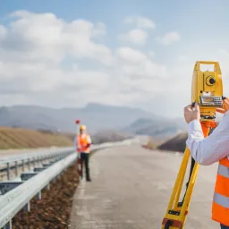 Pomiar geodezyjny nowej drogi: geodeta w pomarańczowej kamizelce i żółtym kasku obsługuje teodolit na tle krajobrazu z błękitnym niebem i chmurami.
