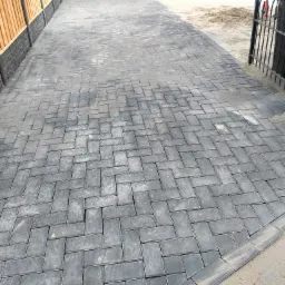 Nowo ułożony podjazd z szarej kostki brukowej w jodełkę, widoczny fragment ogrodzenia i garażu, w tle teren przygotowywany pod dalsze prace.