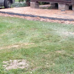 Przygotowanie podłoża pod taras: wyrównany teren z obrzeżami, podsypka z piasku, widoczny fragment konstrukcji tarasu na podwyższeniu i zaparkowany samochód w tle.