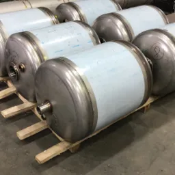 Rząd cylindrycznych zbiorników ze stali nierdzewnej, ustawionych na drewnianej palecie w hali produkcyjnej, z widocznymi spawami i przyłączami.