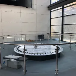 Okrągła szklana bariera z metalowymi słupkami otaczająca interaktywną instalację w nowoczesnym wnętrzu budynku z dużymi oknami i szarą podłogą.