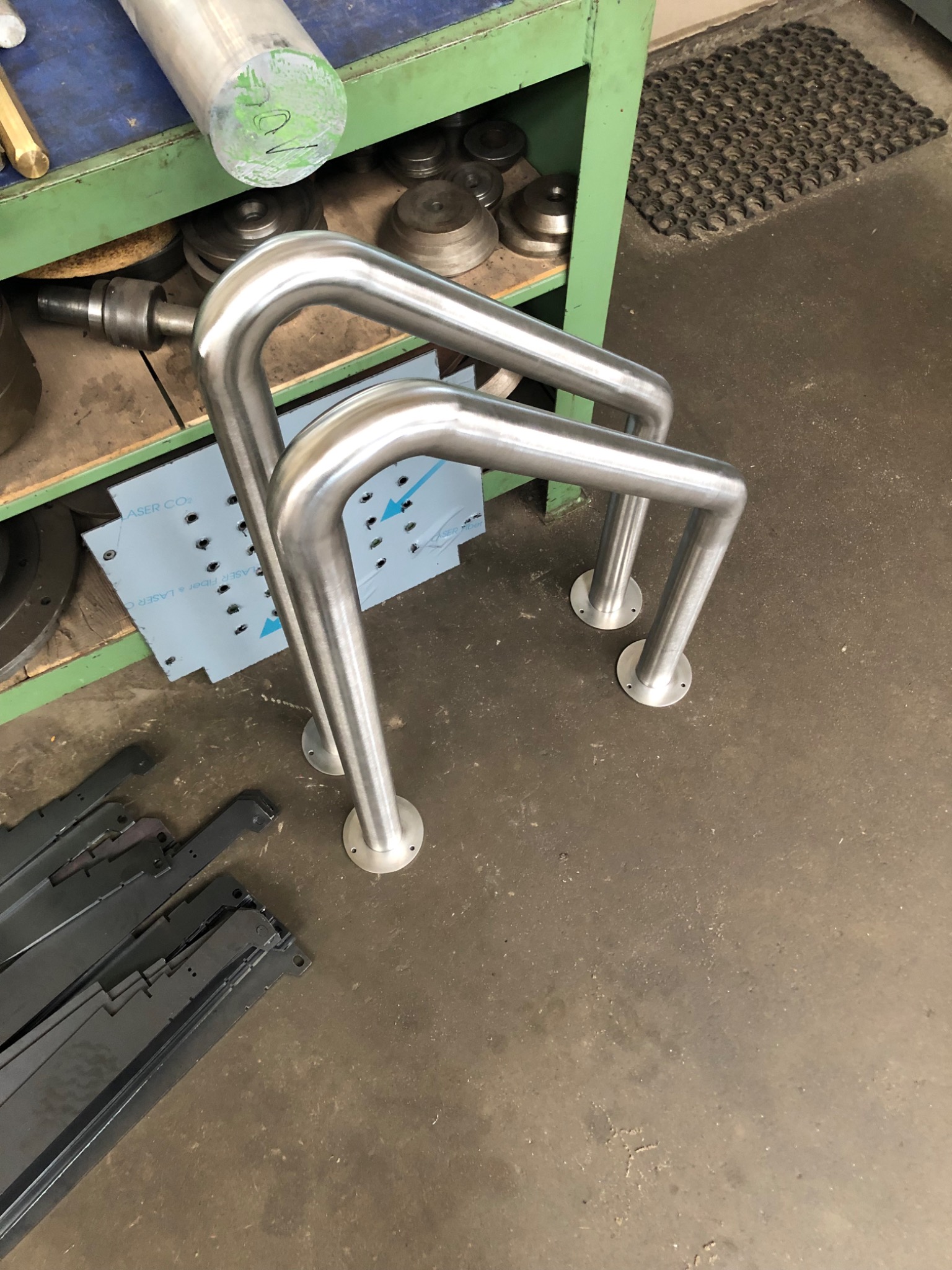 Dwie polerowane poręcze ze stali nierdzewnej z okrągłymi podstawami na tle warsztatu, obok leżą czarne elementy metalowe, a w tle widać narzędzia i fragment arkusza blachy z otworami.