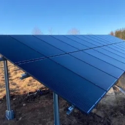 Panel słoneczny zamontowany na metalowej konstrukcji na tle pola z suchą trawą i drzewami w oddali, widoczne przewody.