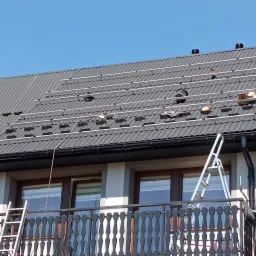 Montaż systemu fotowoltaicznego na dachu z blachodachówki, widoczne szyny montażowe, drabina oparta o balkon, błękitne niebo w tle.