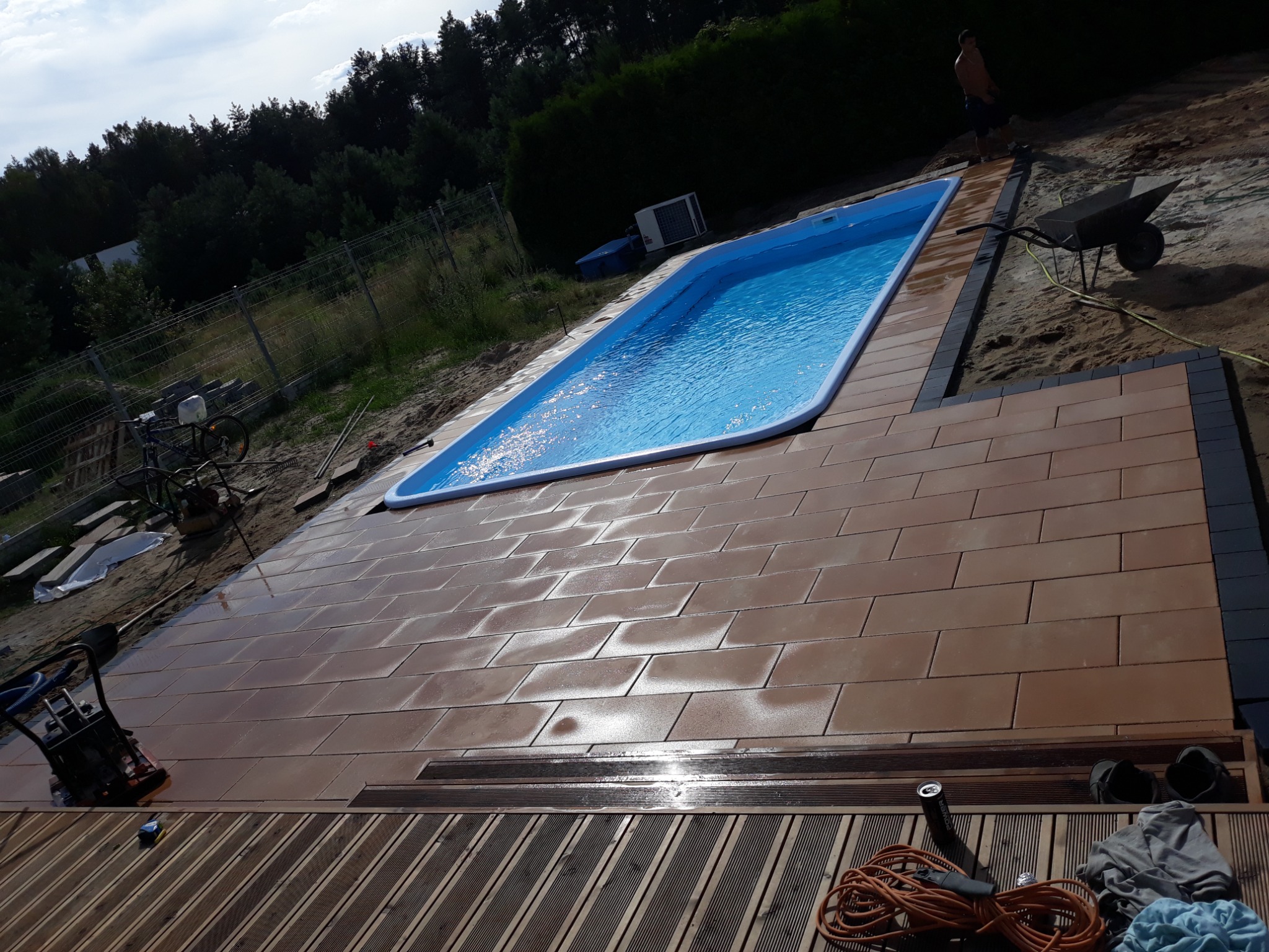 Montaż brązowej kostki brukowej wokół basenu ogrodowego z widocznym ubijakiem wibracyjnym, taczką i fragmentem drewnianego tarasu.