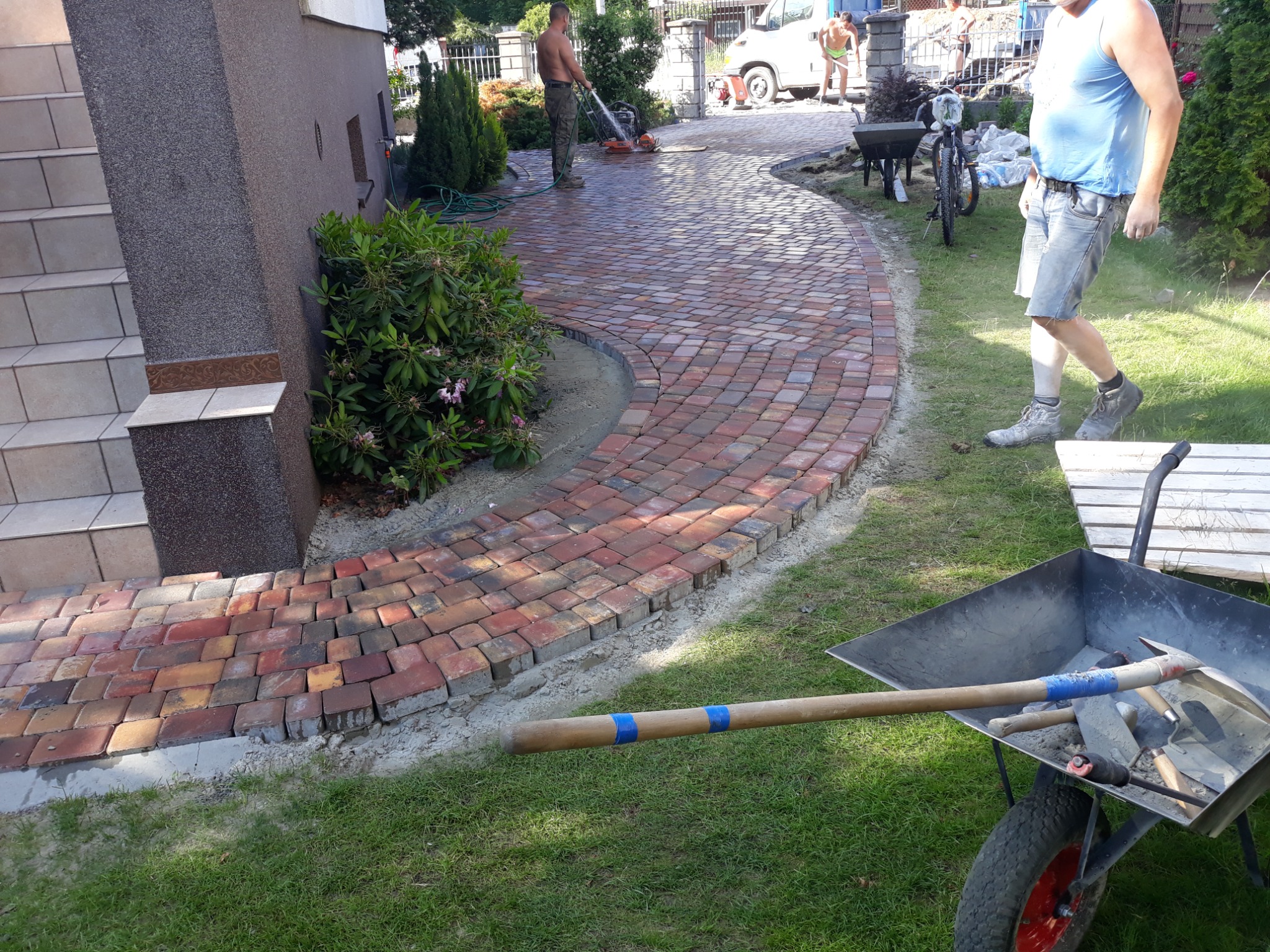 Czerwono-brązowa kostka ułożona w łukowaty chodnik prowadzący od schodów budynku, z pracownikami i narzędziami w tle, wózek budowlany z narzędziami na pierwszym planie.