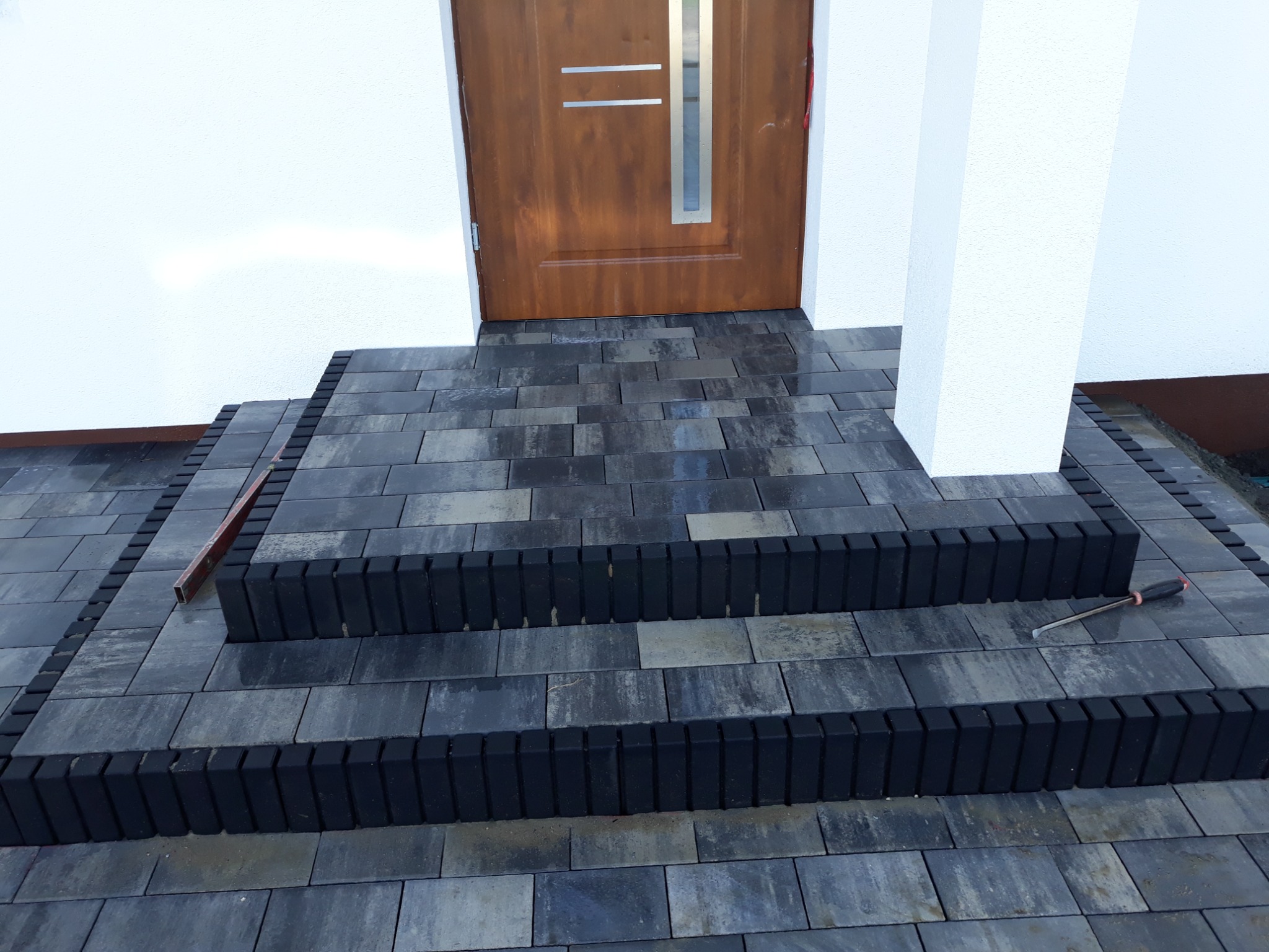 Schody wejściowe do domu wyłożone szarą kostką brukową z czarnymi obrzeżami, widoczny poziomica i dłuto.