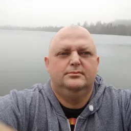 Portret mężczyzny w szarej bluzie z kapturem na tle jeziora spowitego mgłą.