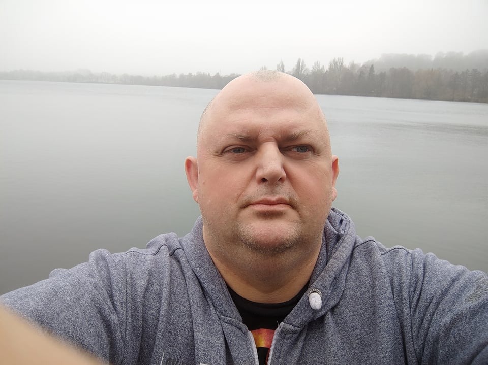 Portret mężczyzny w szarej bluzie z kapturem na tle jeziora spowitego mgłą.