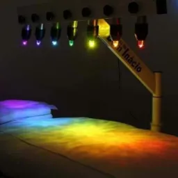 Łóżko do terapii kolorami z lampami emitującymi różne barwy światła, oświetlające materac w gradient tęczy, z widocznym napisem na ramieniu lampy.