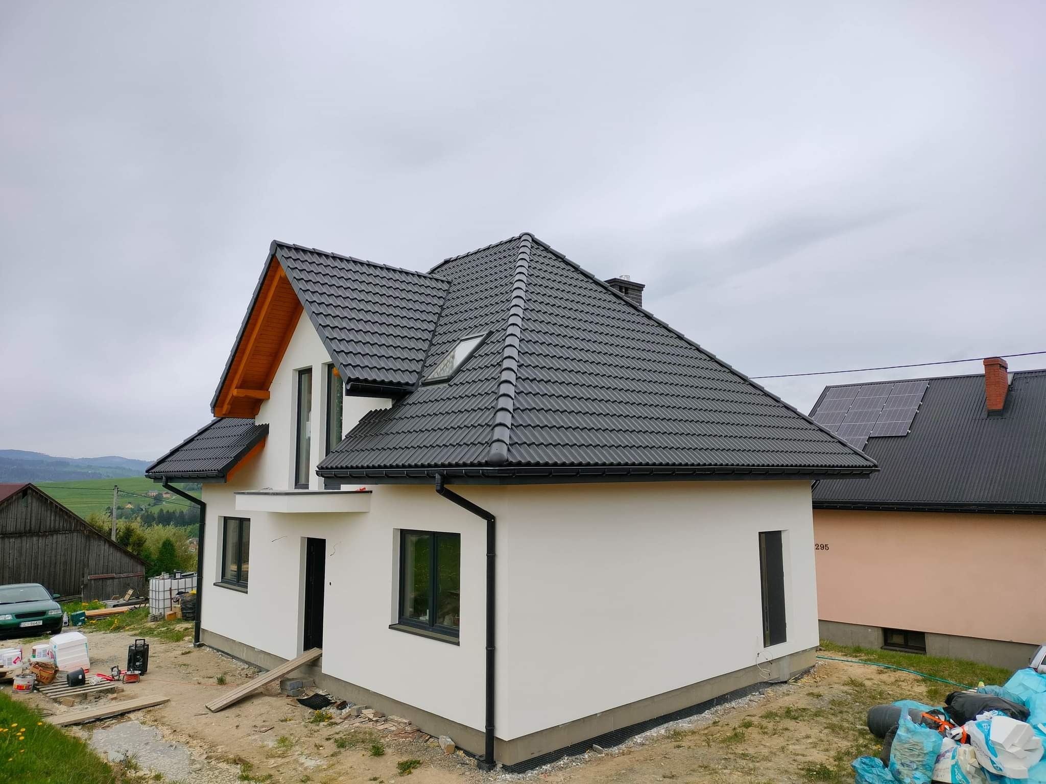 Nowoczesny dom jednorodzinny z białą elewacją i grafitową dachówką, widok z zewnątrz, teren wokół domu w trakcie prac wykończeniowych, widoczne materiały budowlane i narzędzia.