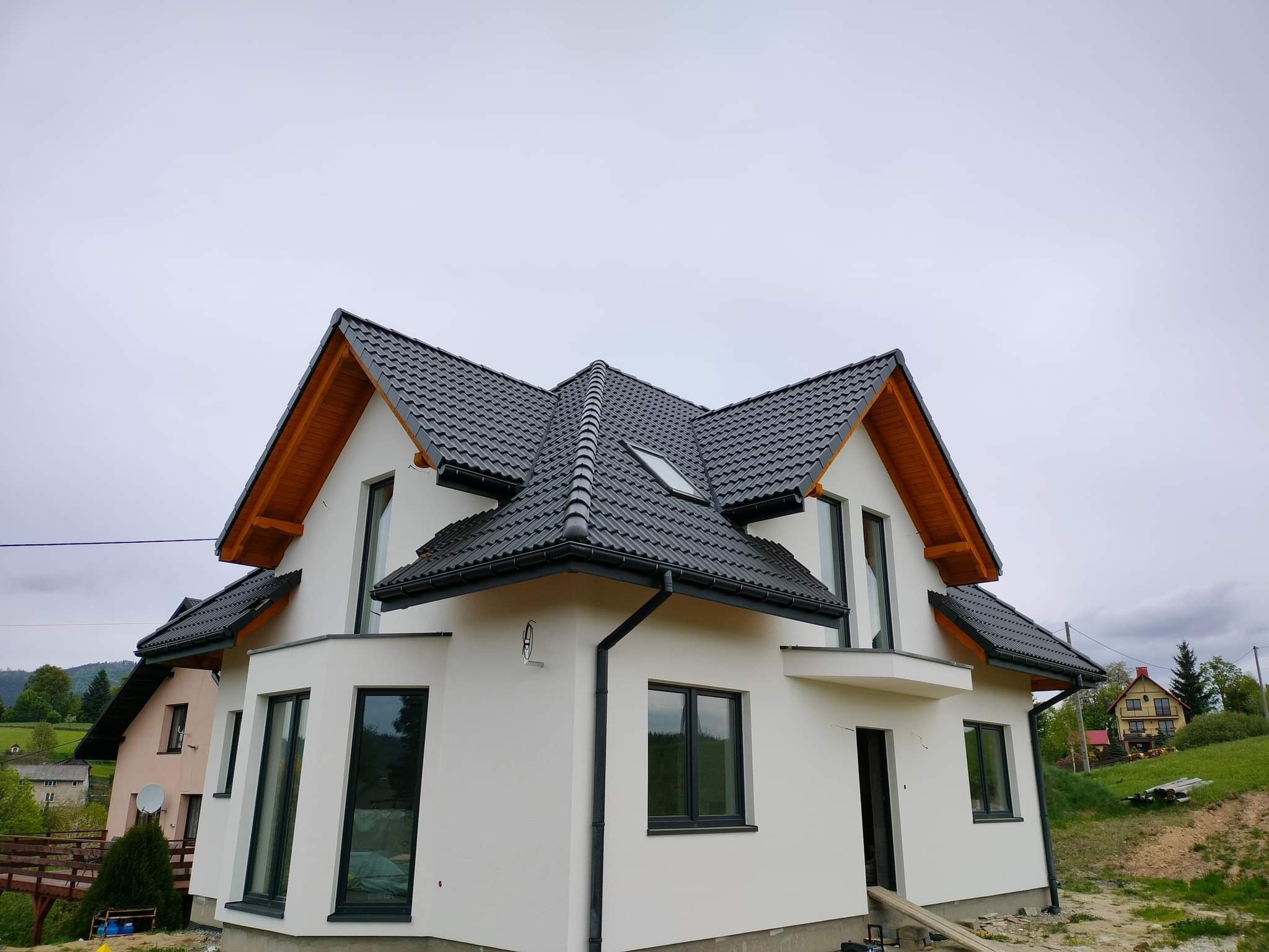Nowoczesny dom jednorodzinny z białą elewacją i czarną dachówką, widok z dołu na skośny dach z oknem dachowym, szare okna i rynny, surowy krajobraz w tle.