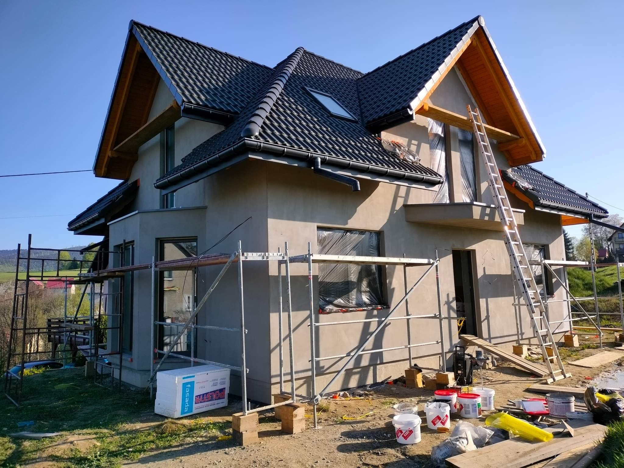 Dom w trakcie prac elewacyjnych: surowa elewacja w szarym kolorze, rusztowania, drabina oparta o okno zabezpieczone folią, puszki z farbą i materiały budowlane na ziemi.