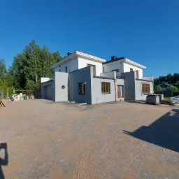 Nowoczesny dom dwukondygnacyjny z szarą elewacją i białymi elementami, widoczny w pełnym słońcu z brukowanego podjazdu z materiałami budowlanymi w tle, cień fotografa w lewym dolnym rogu.