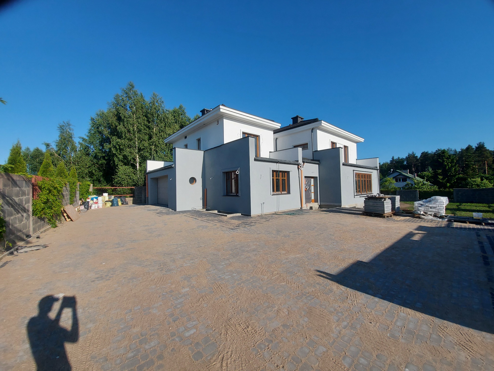 Nowoczesny dom dwukondygnacyjny z szarą elewacją i białymi elementami, widoczny w pełnym słońcu z brukowanego podjazdu z materiałami budowlanymi w tle, cień fotografa w lewym dolnym rogu.