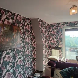 Pokój z tapetą w różowe róże, widoczny fragment sofy, okno i obraz na ścianie.